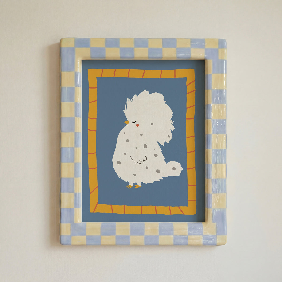 A framed art print featuring a cute white fluffy bird with small gray spots on a dusty blue background, surrounded by a yellow-orange patterned border and a blue-and-beige checkered frame, mounted on a light beige wall.
