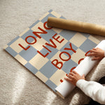 A person unrolling a large poster on a beige carpet. The poster features a light blue and cream checkerboard pattern with the words 'LONG LIVE BOY' printed in bold red letters.