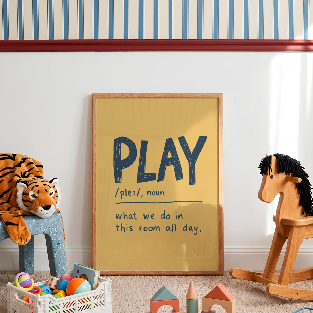 A framed typographic wall art print leaning against a white wall in a styled children's room. The print has a mustard yellow background with the word 'PLAY' in large dark blue letters and a handwritten-style definition below.