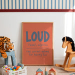 A framed children's wall poster with a terracotta orange background and blue-gray lettering that reads 'LOUD /laʊd/, adjective the correct volume for having fun', styled in a nursery or playroom with a striped wall, toys, a rocking horse, and a stuffed tiger.