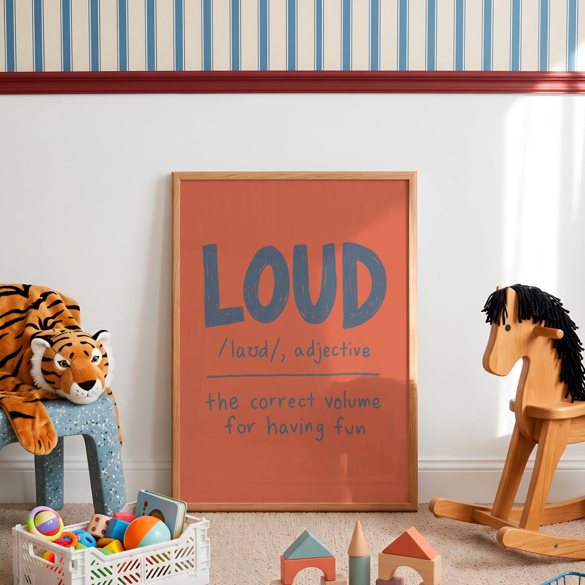 A framed children's wall poster with a terracotta orange background and blue-gray lettering that reads 'LOUD /laʊd/, adjective the correct volume for having fun', styled in a nursery or playroom with a striped wall, toys, a rocking horse, and a stuffed tiger.