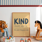A framed children's art print leaning against a white wall in a styled nursery or playroom. The print has a light wood frame and a warm beige background with the word 'KIND' in large dark blue letters, plus pronunciation guidance and the phrase 'the coolest thing you can be.