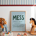A framed typographic wall art print leaning against a white wall in a children's room. The print has a light blue background with dark green text reading 'MESS /mes/, noun proof that something good happened here.