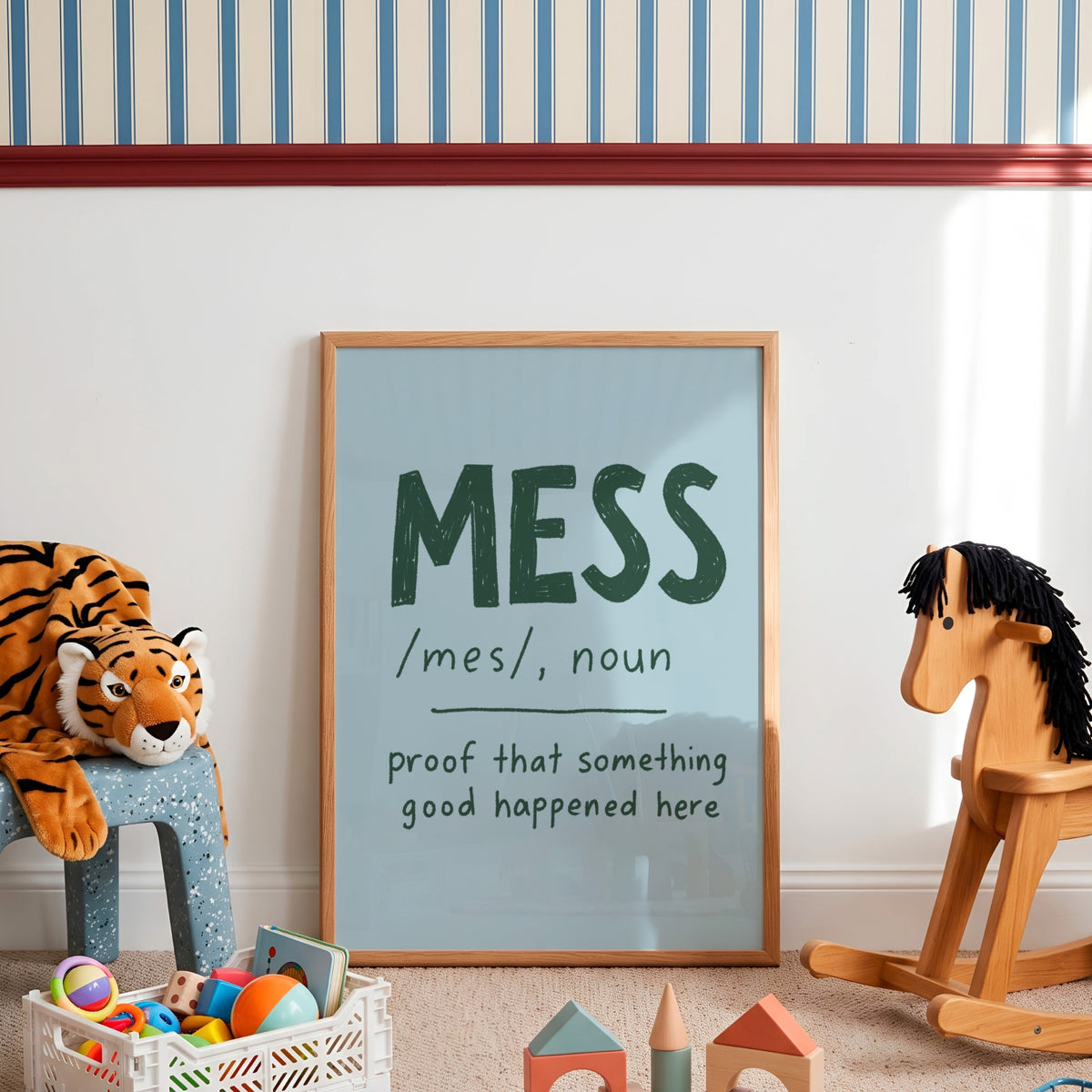 A framed typographic wall art print leaning against a white wall in a children's room. The print has a light blue background with dark green text reading 'MESS /mes/, noun proof that something good happened here.