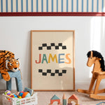 A framed personalized children's art print leaning against a white wall in a styled nursery or playroom. The print features the name 'JAMES' in colorful letters between black checkered stripes on a cream background, enclosed in a light wood frame.