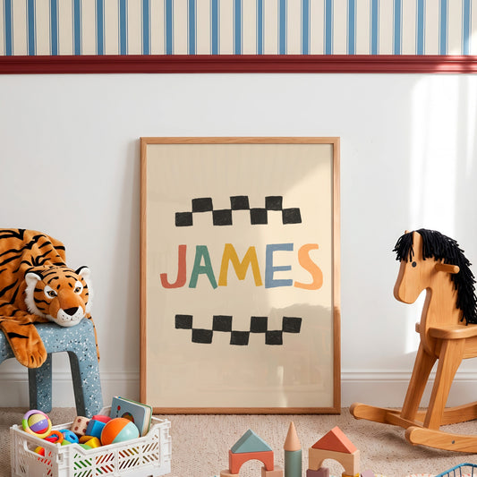 A framed personalized children's art print leaning against a white wall in a styled nursery or playroom. The print features the name 'JAMES' in colorful letters between black checkered stripes on a cream background, enclosed in a light wood frame.