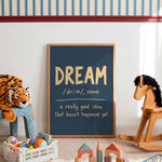 A framed chalkboard-style wall art print leaning against a white wall in a styled children's room. The print reads 'DREAM /dri:m/, noun' with the definition 'a really good idea that hasn't happened yet' in gold and white lettering on a dark blue background.