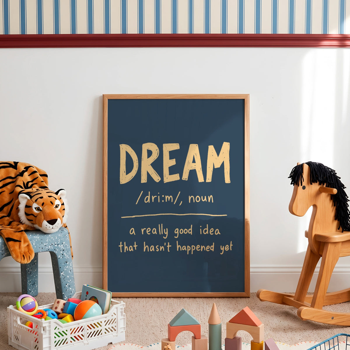 A framed chalkboard-style wall art print leaning against a white wall in a styled children's room. The print reads 'DREAM /dri:m/, noun' with the definition 'a really good idea that hasn't happened yet' in gold and white lettering on a dark blue background.
