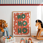 A framed art print featuring the words 'HO HO HO' in green on an orange background with white polka dots, styled in a child's playroom next to a wooden rocking horse and toys.