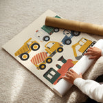 A child's nursery wall mural featuring colorful construction vehicles, shown partially unrolled on a beige carpet with a cardboard roll and hands visible.
