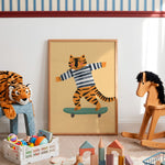 A framed art print featuring a cartoon tiger wearing a striped shirt and balancing on a skateboard, displayed in a child's playroom next to a stuffed tiger and a wooden rocking horse.