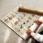 A safari-themed alphabet mat being rolled out on a beige carpet, with a person's hands visible and a brown cardboard shipping tube nearby.