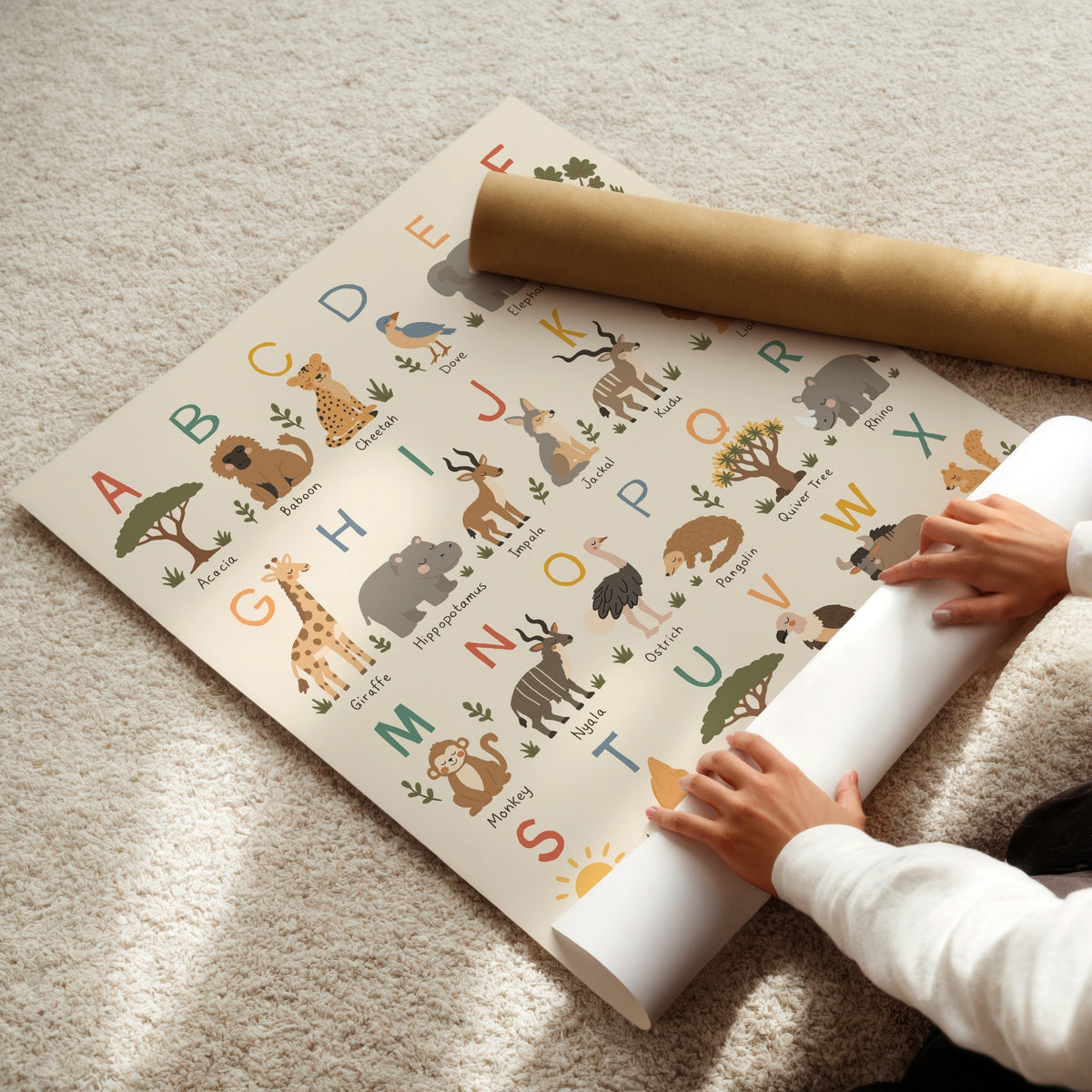 A safari-themed alphabet mat being rolled out on a beige carpet, with a person's hands visible and a brown cardboard shipping tube nearby.
