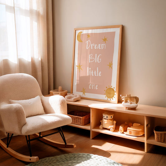 A framed nursery wall art print with the text 'Dream Big little one' in white script, decorated with gold stars and a crescent moon on a dusty pink background, leaning on a light wood shelf in a sunlit nursery next to a cream boucle rocking chair.