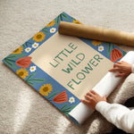 A person unrolling a decorative poster or mat that reads “LITTLE WILD FLOWER,” with a matching rolled sheet and a brown paper roll on a beige carpet. The design has a blue border with yellow sunflowers, white daisies, and coral flower shapes.