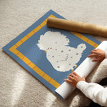 A person is unrolling a large decorative wall mural or poster featuring a cute white speckled chick on a blue background with a yellow border, placed on a beige carpet. A brown cardboard mailing tube is also visible.
