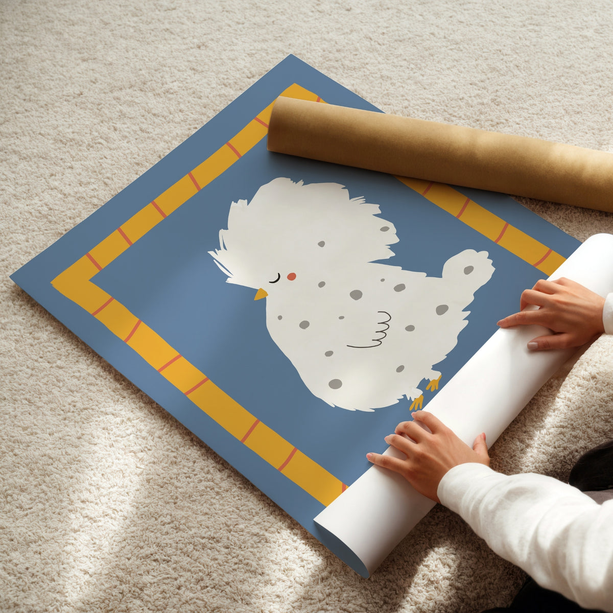 A person is unrolling a large decorative wall mural or poster featuring a cute white speckled chick on a blue background with a yellow border, placed on a beige carpet. A brown cardboard mailing tube is also visible.