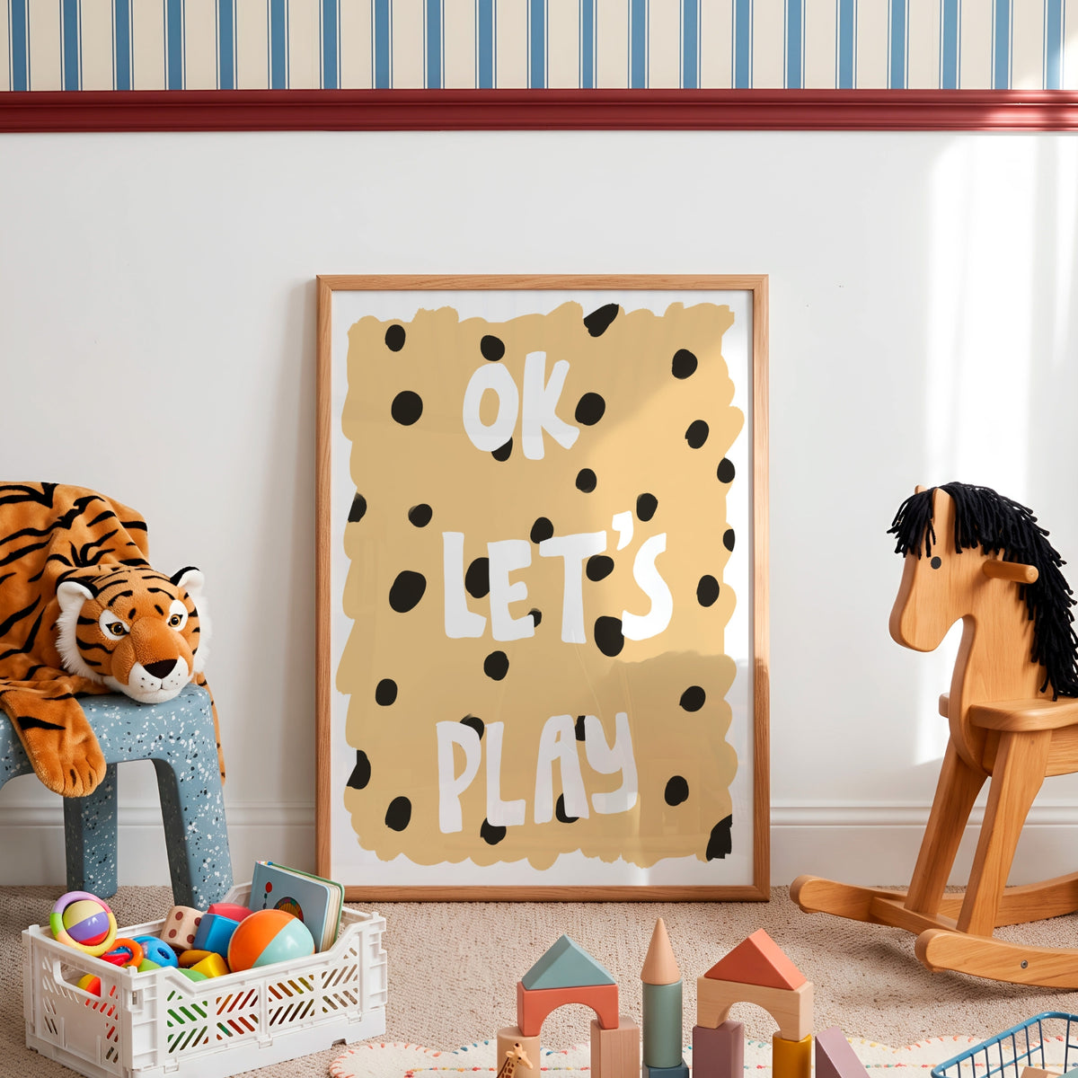 A framed children's wall art print reading 'OK LET'S PLAY' in white letters on a tan background with black spots, styled in a nursery or playroom with toys, a tiger plush, and a wooden rocking horse.