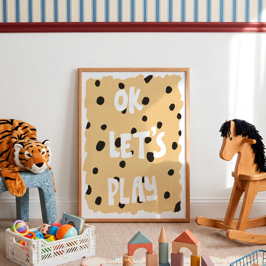 A framed children's wall art print reading 'OK LET'S PLAY' in white letters on a tan background with black spots, styled in a nursery or playroom with toys, a tiger plush, and a wooden rocking horse.