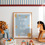 A framed nursery art print leaning against a white wall in a styled children's room. The print has a light blue background with white text reading 'Dream BIG little one' and gold star and sun illustrations.
