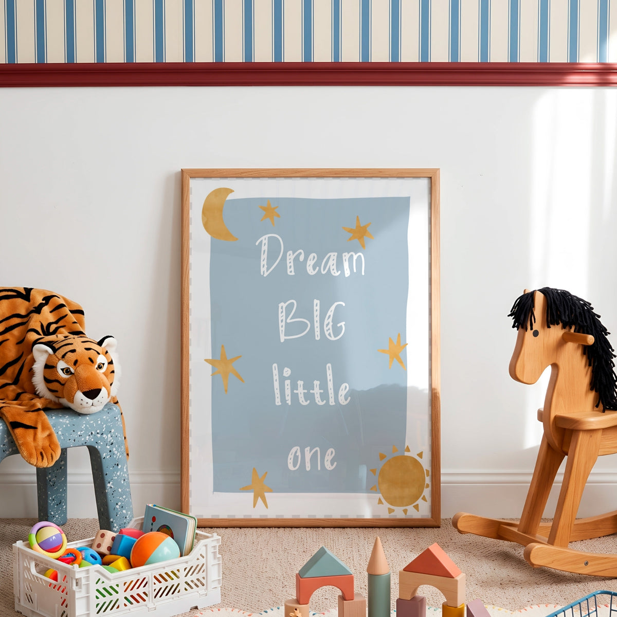 A framed nursery art print leaning against a white wall in a styled children's room. The print has a light blue background with white text reading 'Dream BIG little one' and gold star and sun illustrations.