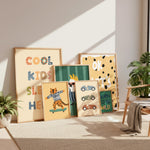 A styled collection of seven framed kids' art prints with various colorful illustrations and text, arranged on a beige rug in a sunlit room next to a wooden chair and potted plants.