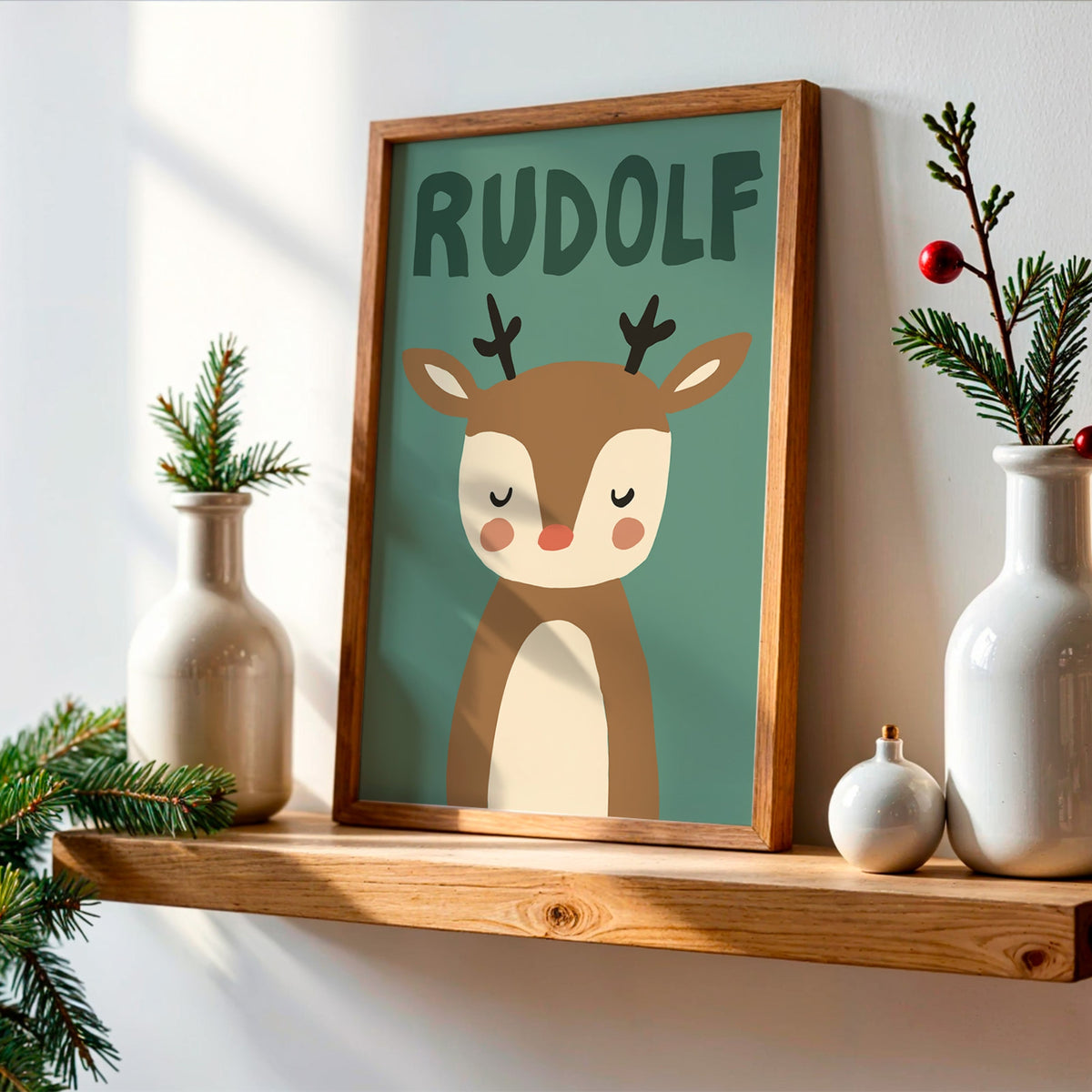 A framed personalized art print featuring a cute cartoon reindeer with the name 'RUDOLF' above it, styled on a wooden shelf with white ceramic vases and pine branches.