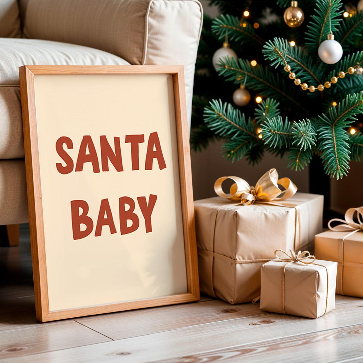 A framed Christmas sign leaning against a beige sofa on a wooden floor, next to wrapped gift boxes and a decorated Christmas tree. The sign has a light wood frame, a cream background, and the words 'SANTA BABY' in large reddish-brown letters.