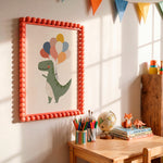 A framed children's wall art print hangs on a sunlit wall above a wooden desk. The print shows a green dinosaur holding colorful balloons, surrounded by a bright coral bead frame.