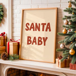 A framed art print with the text 'SANTA BABY' in a warm reddish-brown font on a cream background, styled on a white fireplace mantel next to a decorated Christmas tree and wrapped gifts.