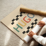 A person unrolls a large beige wall poster or wallpaper sheet on a light beige carpet. The design features the word 'HUGO' in colorful retro letters (green, red, yellow, blue) framed by black checkerboard patterns.