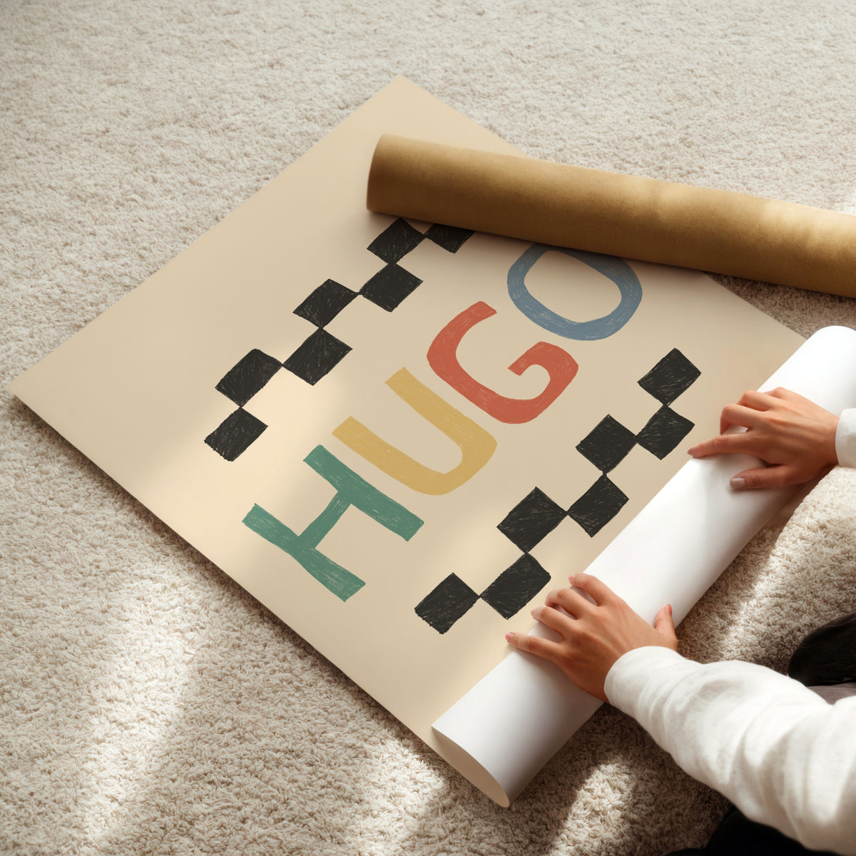 A person unrolls a large beige wall poster or wallpaper sheet on a light beige carpet. The design features the word 'HUGO' in colorful retro letters (green, red, yellow, blue) framed by black checkerboard patterns.