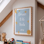 A framed nursery wall art print with the text 'Dream BIG little one' in white lettering on a light blue background, surrounded by yellow stars, a crescent moon, and a sun. The print is housed in a scalloped light wood frame and displayed on a white wall in a child's room with toys and a macrame hanging visible.
