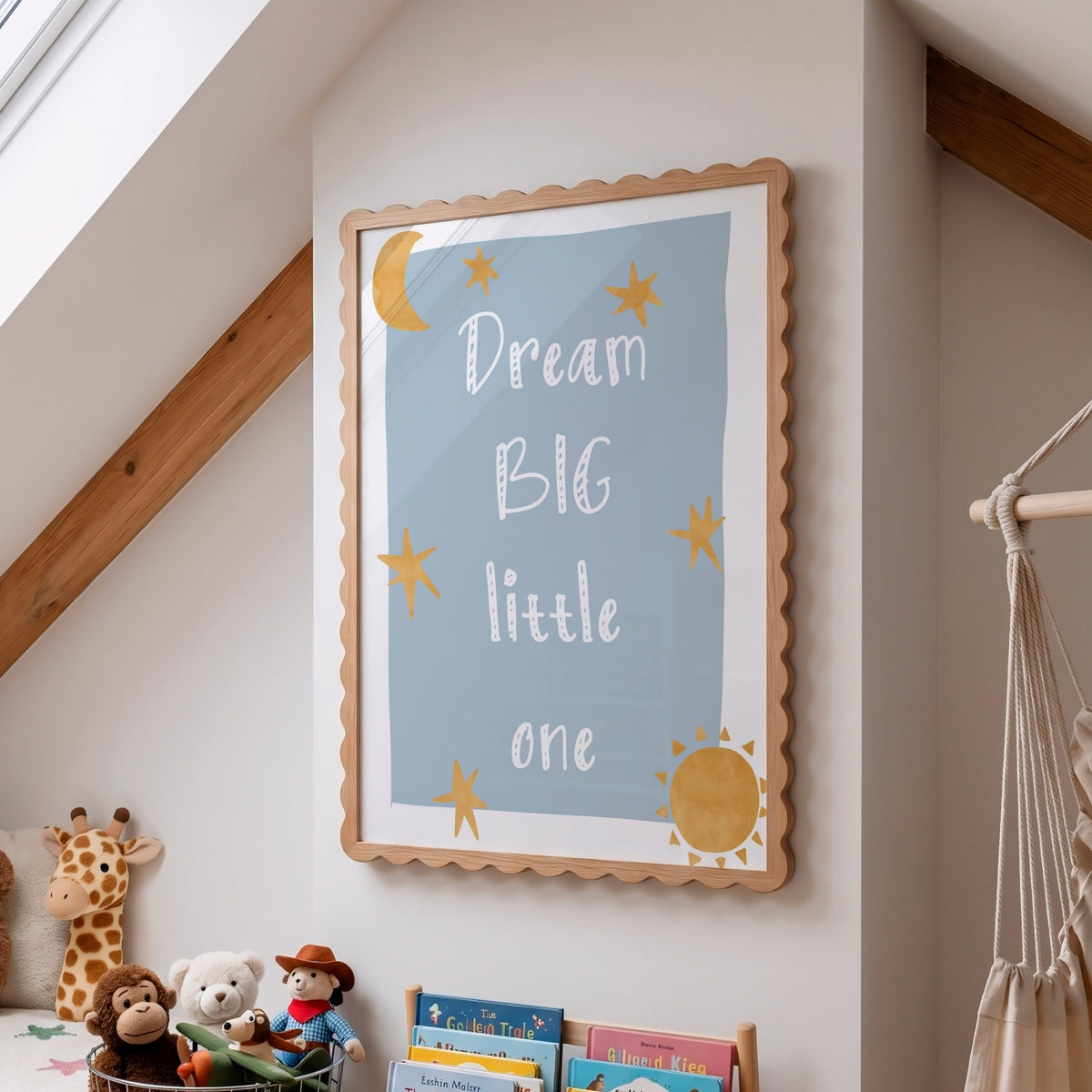 A framed nursery wall art print with the text 'Dream BIG little one' in white lettering on a light blue background, surrounded by yellow stars, a crescent moon, and a sun. The print is housed in a scalloped light wood frame and displayed on a white wall in a child's room with toys and a macrame hanging visible.