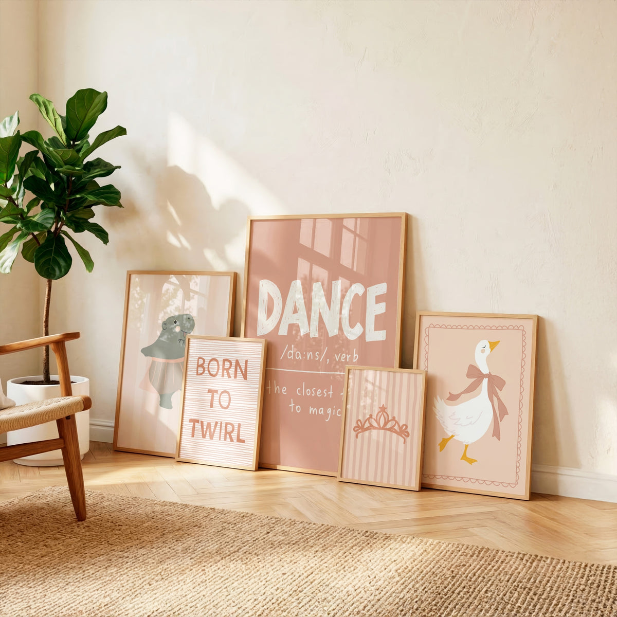 A set of five framed art prints with pastel pink and beige tones arranged on a light wood floor against a cream wall, next to a potted fiddle-leaf fig plant. The prints feature whimsical illustrations and text including 'DANCE', 'BORN TO TWIRL', and a duck with a tiara.