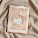 A framed art print featuring a white duck with a brown bow tie on a soft pink background, styled on a textured beige knit blanket.