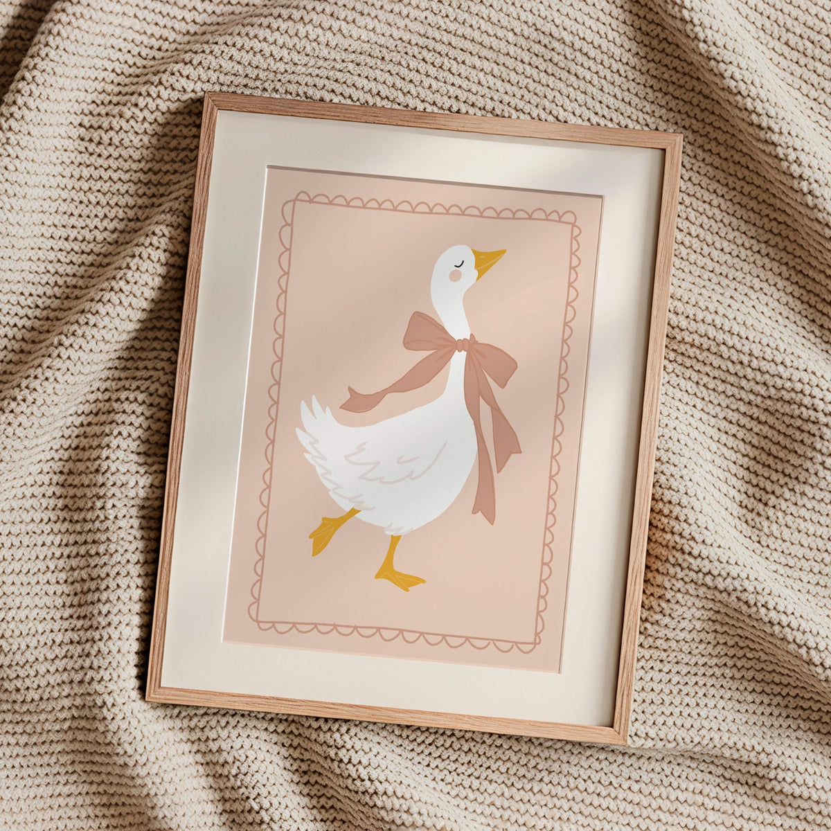 A framed art print featuring a white duck with a brown bow tie on a soft pink background, styled on a textured beige knit blanket.