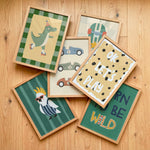 Six framed children's art prints arranged in a flat-lay on a light wood floor. The prints feature whimsical illustrations including a green dinosaur roller skate, blue and gray race cars, a white bird with sunglasses, a green crocodile, and text reading 'OK LET'S PLAY' and 'CARN BE WILD'.