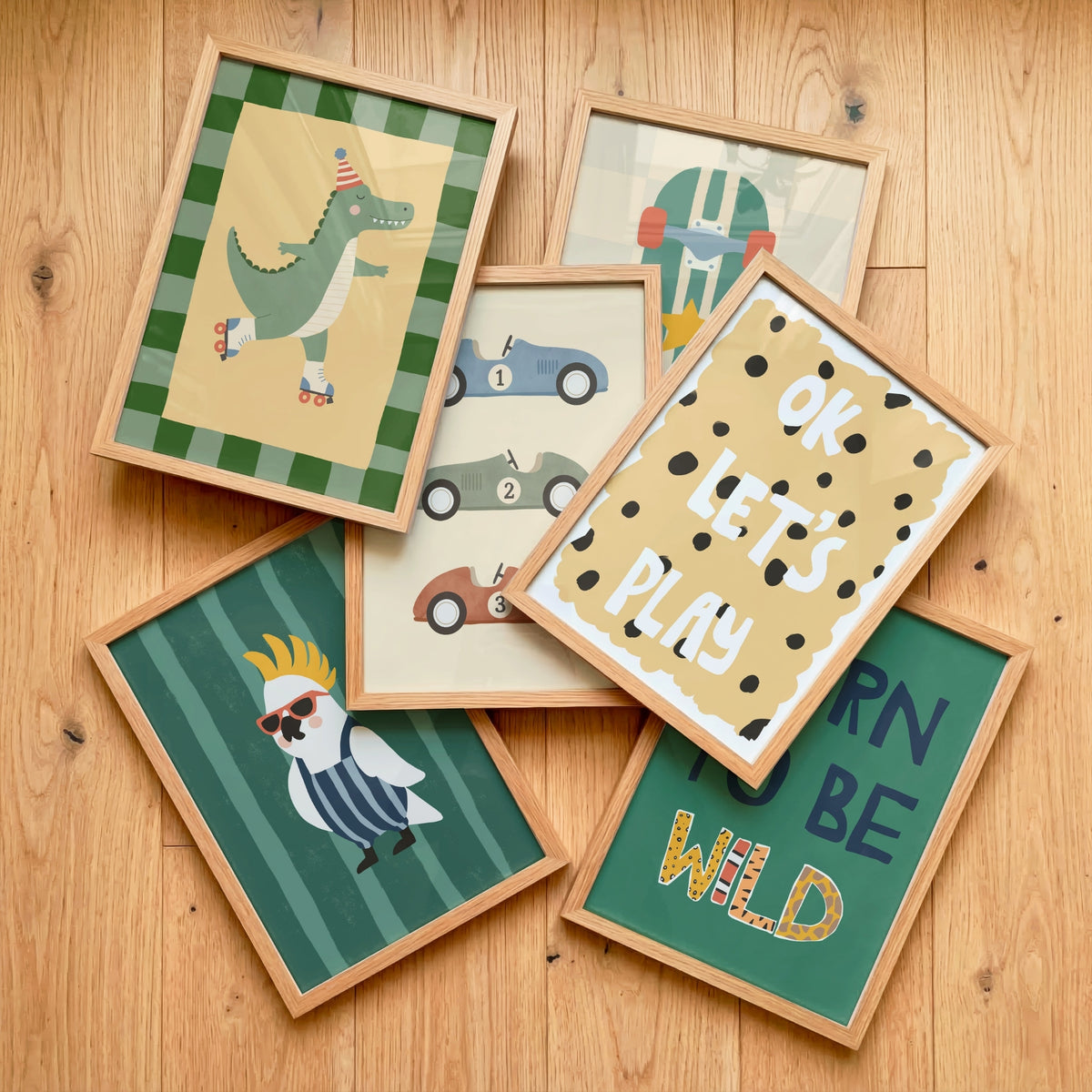 Six framed children's art prints arranged in a flat-lay on a light wood floor. The prints feature whimsical illustrations including a green dinosaur roller skate, blue and gray race cars, a white bird with sunglasses, a green crocodile, and text reading 'OK LET'S PLAY' and 'CARN BE WILD'.