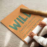 A person unrolling a large orange poster on a beige carpet, with a brown cardboard mailing tube placed on top. The poster features the word 'WILD' in large green letters with a dictionary-style definition underneath.