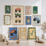 A styled children's nursery wall display featuring multiple framed monster truck and dinosaur-themed art prints in warm wood frames, arranged above a light wood floor with pillows, a stuffed bunny, and a small toy.