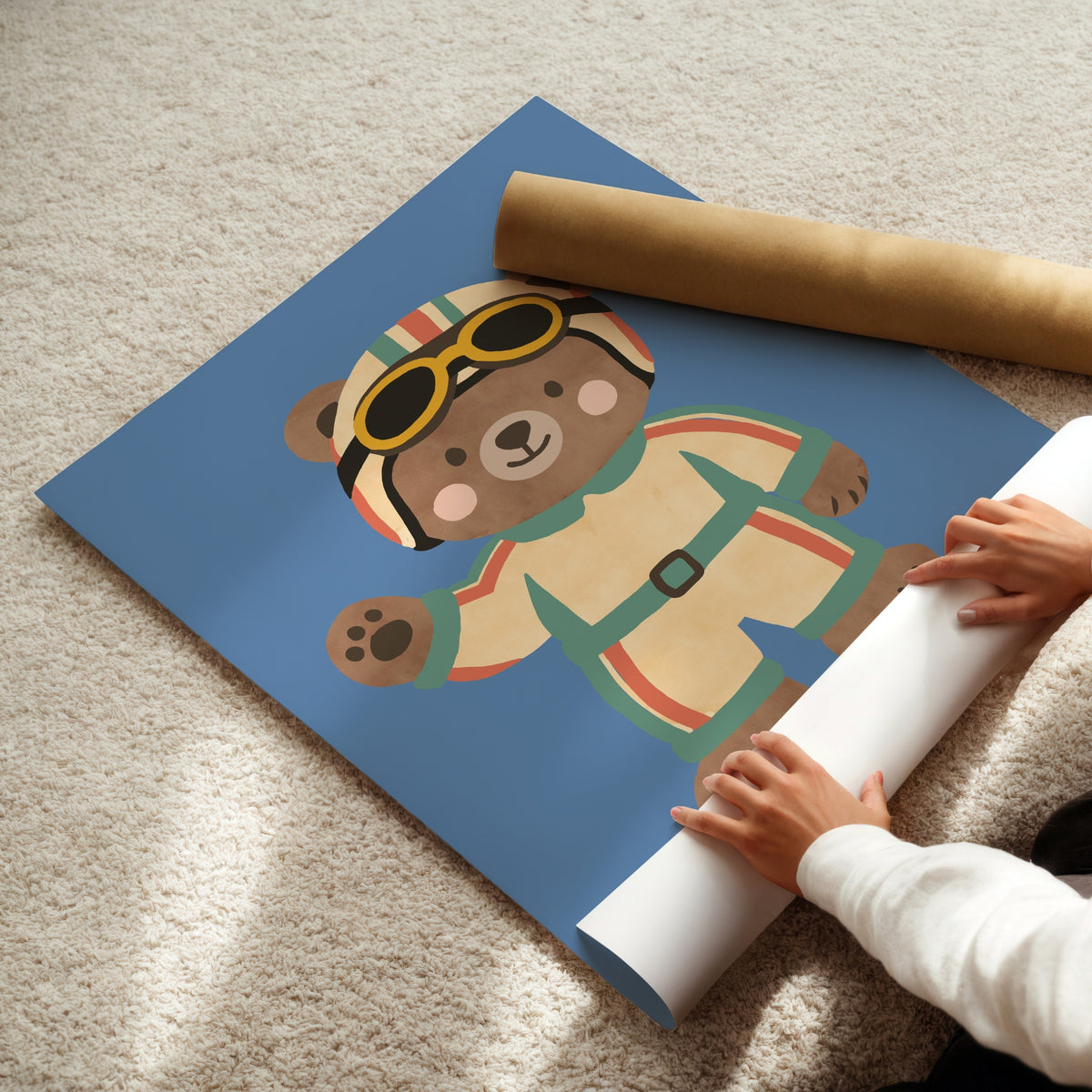 Hands unrolling racing bear art print from kraft shipping tube, showing teddy in race suit and goggles on blue