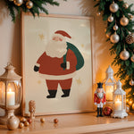A framed Christmas art print featuring a cartoon Santa Claus with a green sack, displayed on a wooden mantel with warm holiday decor including lanterns, ornaments, pine branches, and a nutcracker.