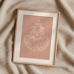 A framed minimalist art print featuring a white line drawing of a ballerina's tutu on a dusty pink background, styled on a textured beige knit blanket.