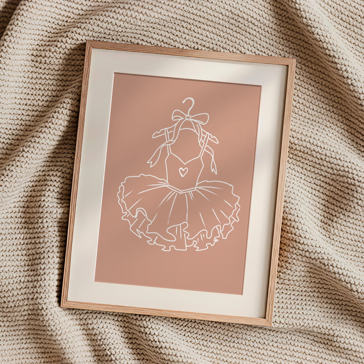 A framed minimalist art print featuring a white line drawing of a ballerina's tutu on a dusty pink background, styled on a textured beige knit blanket.
