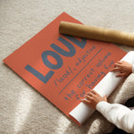A person unrolls a large orange poster on a beige carpet, revealing blue text that reads 'LOUD /laʊd/, adjective the correct volume for having fun.' A brown cardboard mailing tube lies across the poster.