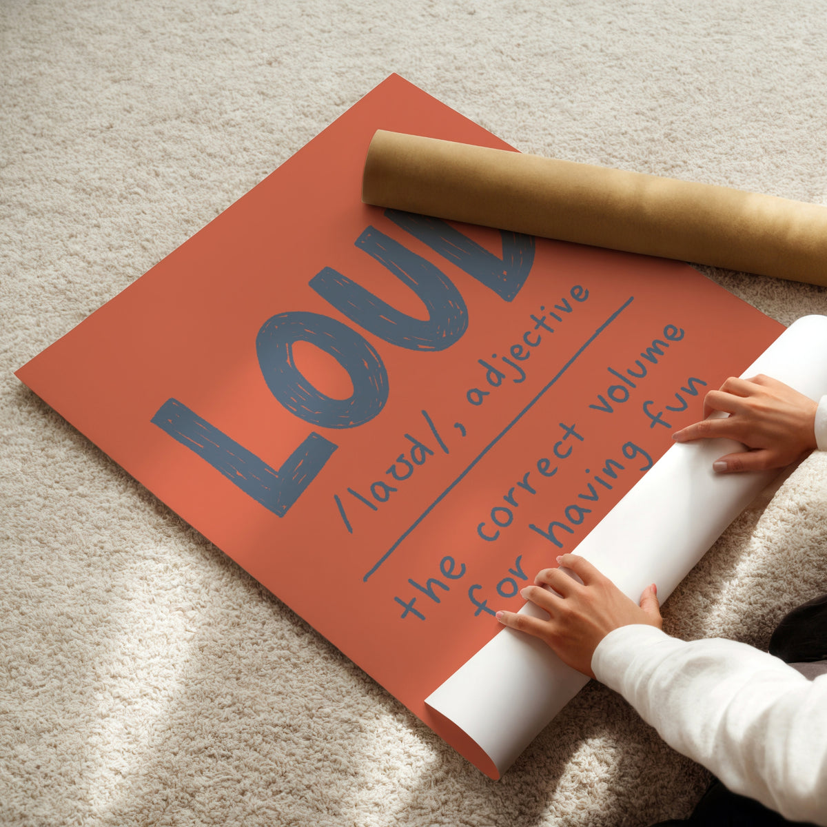 A person unrolls a large orange poster on a beige carpet, revealing blue text that reads 'LOUD /laʊd/, adjective the correct volume for having fun.' A brown cardboard mailing tube lies across the poster.
