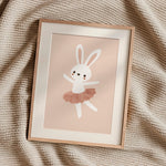 A framed art print featuring a cute white bunny ballerina with a dusty pink tutu, styled on a textured beige knit blanket.