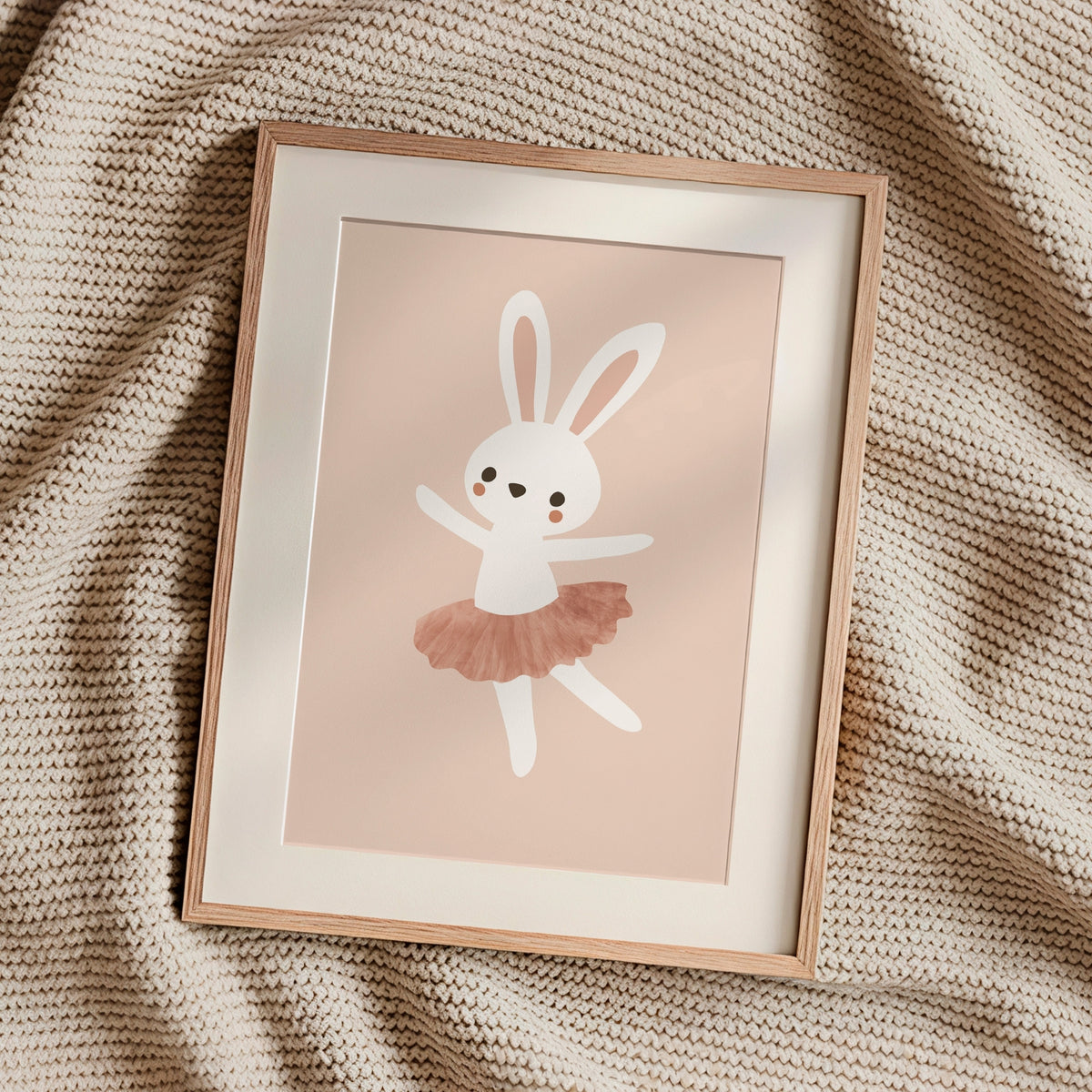 A framed art print featuring a cute white bunny ballerina with a dusty pink tutu, styled on a textured beige knit blanket.