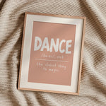 A framed typographic art print reading “DANCE /dæns/, verb” and “the closest thing to magic.” The print has a dusty pink background with white lettering, set in a light wood frame on a textured beige knit blanket.